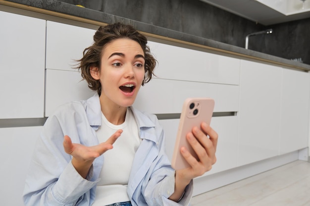 Portrait d'une jeune femme étonnée et sincère tenant un smartphone réagit surprise à de grandes nouvelles conversations vidéo avec quelqu'un assis sur le sol de la cuisine
