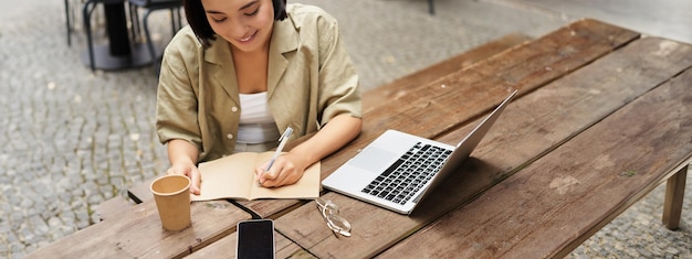 Portrait d'une jeune femme étudiant en ligne assise avec un ordinateur portable, écrivant, prenant des notes et regardant