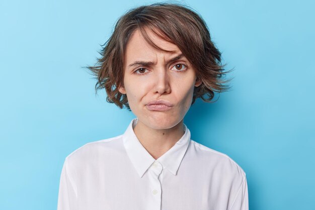 Portrait d'une jeune femme européenne mécontente avec de courts cheveux noirs fronce les sourcils le visage a une expression agacée réagit sur quelque chose de désagréable porte une chemise blanche formelle isolée sur fond de studio bleu