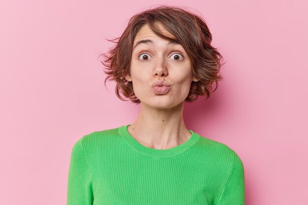 Portrait d'une jeune femme européenne romantique garde les lèvres arrondies veut vous embrasser a une coiffure à la mode porte un pull vert décontracté isolé sur fond de studio rose. Concept d'expressions de visage humain