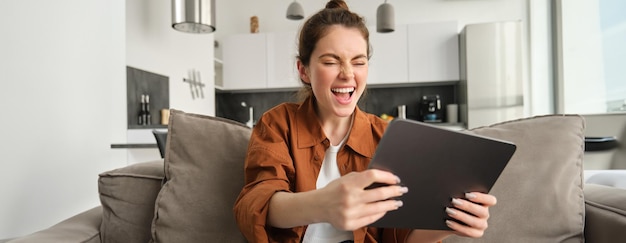 Photo gratuite portrait d'une jeune femme excitée avec une tablette numérique assise sur le canapé en train de rire et de sourire