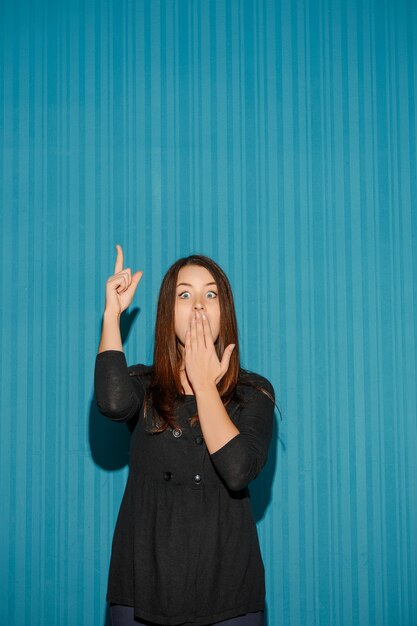 Portrait de jeune femme avec une expression faciale choquée sur fond bleu studio pointant vers le haut