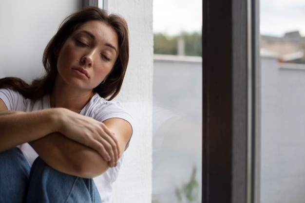 Photo gratuite portrait de jeune femme avec une faible estime de soi assise près de la fenêtre à la maison