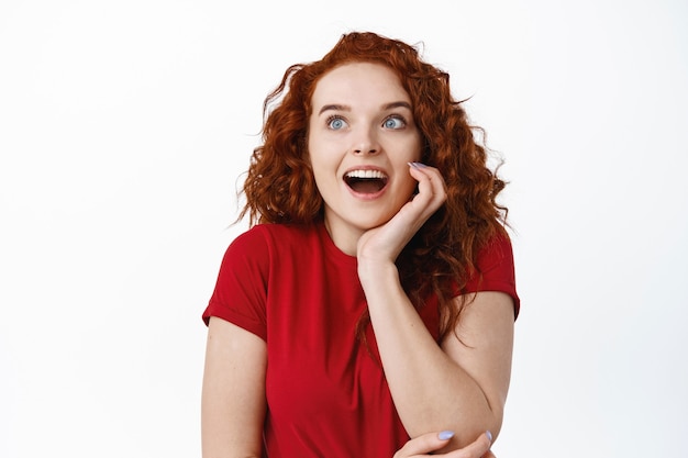 Portrait d'une jeune femme fascinée avec un hiar bouclé rouge, haletant étonné, regardant excité sur le côté gauche avec un espace de copie debout sur un mur blanc