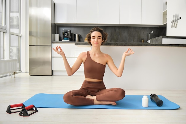 Photo gratuite portrait de jeune femme fitness athlète féminine pratique le yoga à la maison assis en posture de lotus avec re