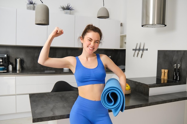 Photo gratuite portrait d'une jeune femme en forme debout dans le salon tenant un tapis de yoga en caoutchouc flexant ses biceps.