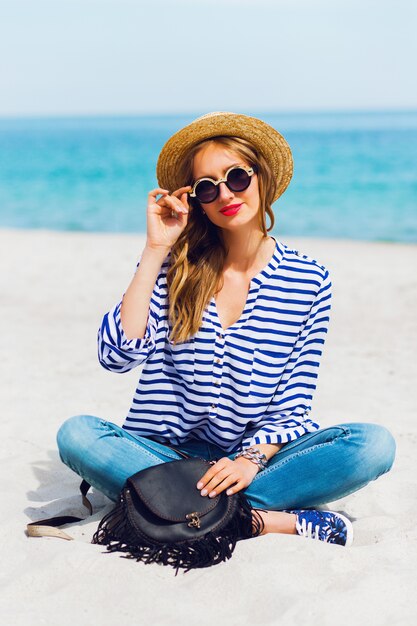 Portrait de jeune femme fraîche dans des lunettes de soleil cool et chapeau de paille assis sur la plage tropicale ensoleillée