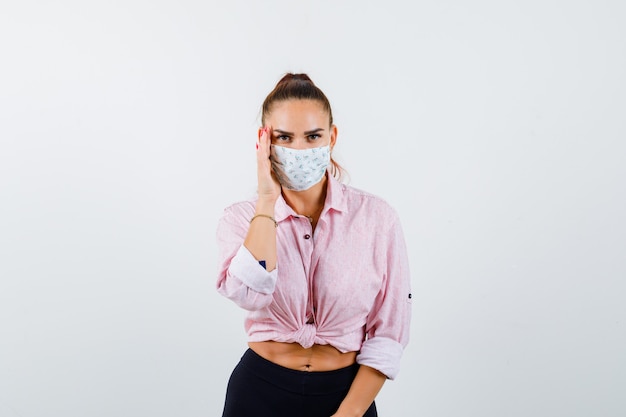 Portrait De Jeune Femme En Gardant La Main Sur Le Côté Du Visage En Chemise, Pantalon, Masque Et à La Vue De Face Curieuse