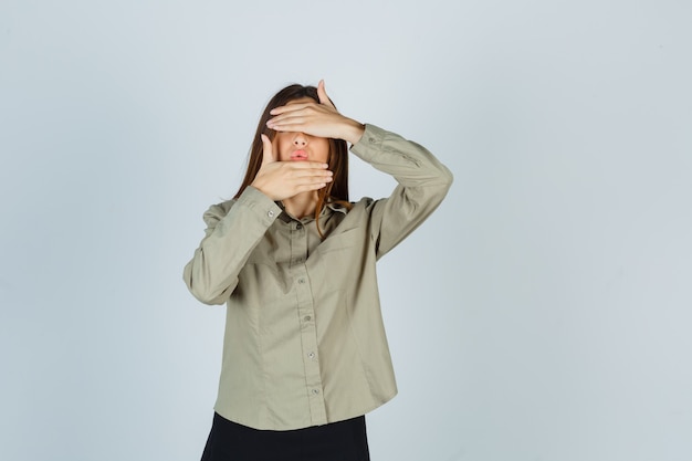 Portrait de jeune femme gardant la main sur les yeux et le menton, faisant la moue des lèvres en chemise, jupe et regardant effrayé vue de face