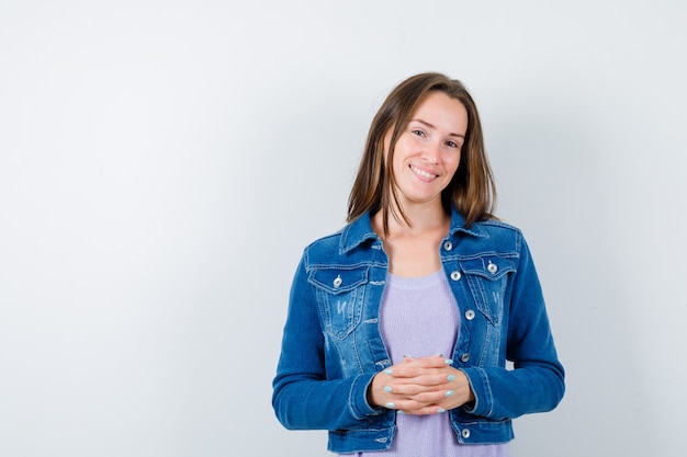 Portrait de jeune femme gardant les mains jointes devant elle en t-shirt, veste et à la vue de face joyeuse