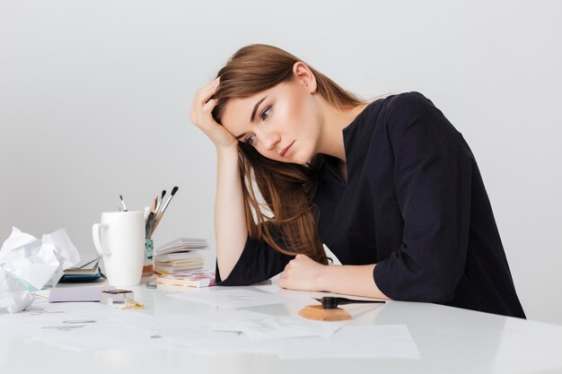 Portrait de jeune femme gentille assise au bureau blanc et se penchant la tête sur sa main tout en regardant de côté fatigué isolé