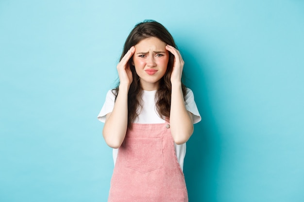 Portrait de jeune femme grimaçant de migraine douloureuse, fronçant les sourcils et touchant la tête, ayant des maux de tête, debout sur fond bleu.