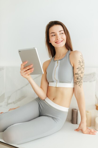 Portrait de jeune femme en haut sportif et leggings assis sur une table de cuisine avec tablette à la main et regardant joyeusement de côté