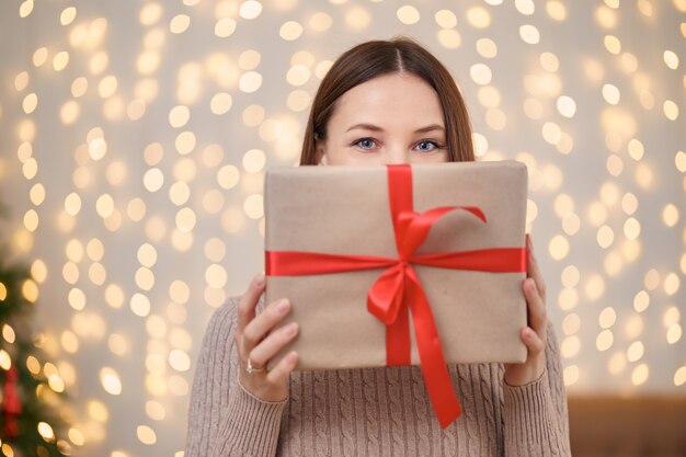 Portrait de jeune femme heureuse lèvres rouges se cachant derrière une boîte cadeau enveloppée.