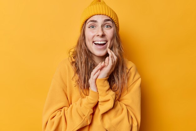 Portrait d'une jeune femme heureuse sourit à pleines dents a surpris l'expression des dents blanches parfaites rit réagit joyeusement sur des nouvelles impressionnantes porte un sweat-shirt décontracté et un chapeau isolé sur fond jaune