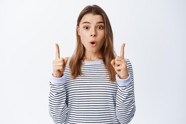 Portrait d'une jeune femme impressionnée vérifiant une publicité cool, pointant les doigts vers le haut et disant wow, montrant le chemin, debout sur blanc