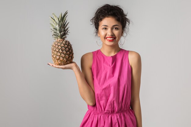 Portrait de jeune femme jolie positive en robe rose, tenant l'ananas, émotion drôle, heureux, souriant, style d'été, régime de fruits, regardant à huis clos, pensée, asiatique, métisse, isolé