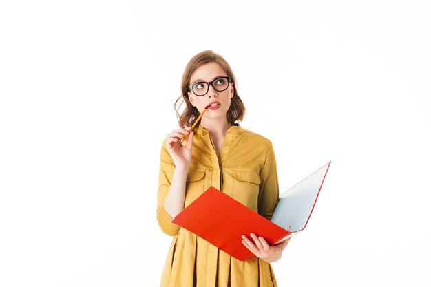 Portrait de jeune femme à lunettes debout avec dossier rouge ouvert et crayon dans les mains et regardant rêveusement de côté sur fond blanc isolé