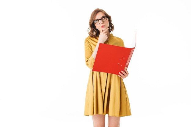 Portrait de jeune femme à lunettes et robe jaune debout avec dossier rouge à la main et regardant pensivement de côté sur fond blanc isolé