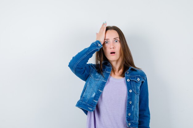 Portrait de jeune femme avec la main sur la tête en t-shirt, veste et à la vue de face choquée