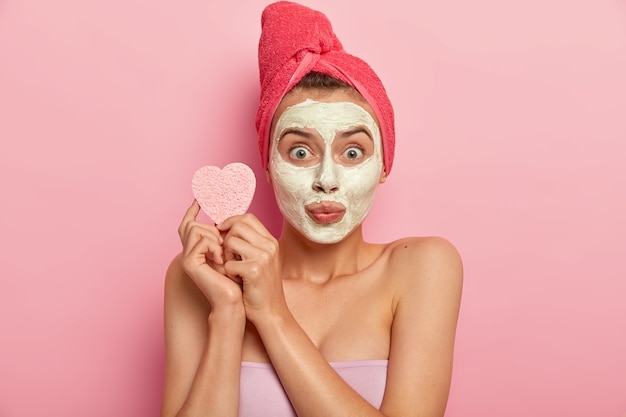 Portrait de jeune femme avec un masque naturel minéral sur le visage, a un traitement quotidien de soins de la peau, tient une éponge cosmétique pour essuyer le teint, purifie la peau et élimine les cellules mortes de la peau, a un bain à la maison