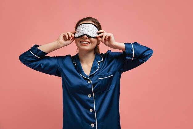 Portrait de jeune femme en masque de sommeil et pyjama bleu posant isolé sur fond de studio rose