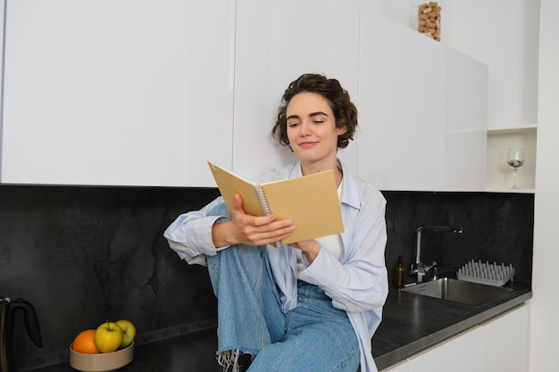 Portrait d'une jeune femme moderne lisant dans la cuisine, assise sur le comptoir et souriante étudiant