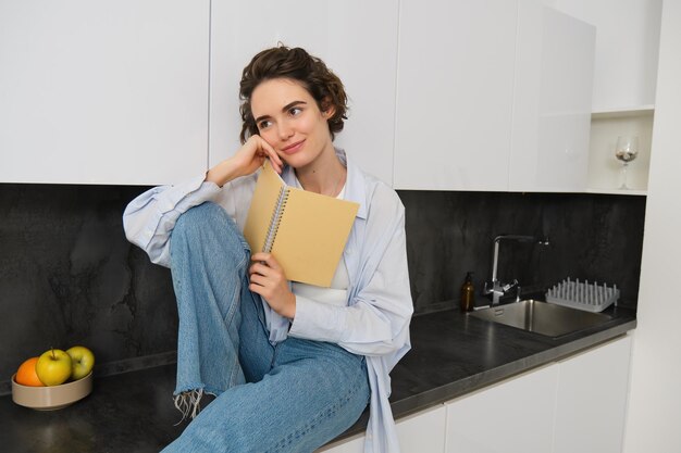 Portrait d'une jeune femme moderne lisant dans la cuisine, assise sur le comptoir et souriante étudiant