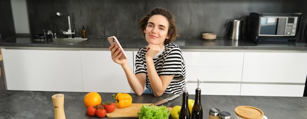 Photo gratuite portrait d'une jeune femme moderne qui réfléchit à ce qu'elle doit cuisiner assise dans la cuisine avec un smartphone et