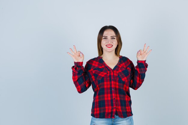 Portrait de jeune femme montrant un geste correct dans une chemise à carreaux, un jean et l'air heureux en vue de face