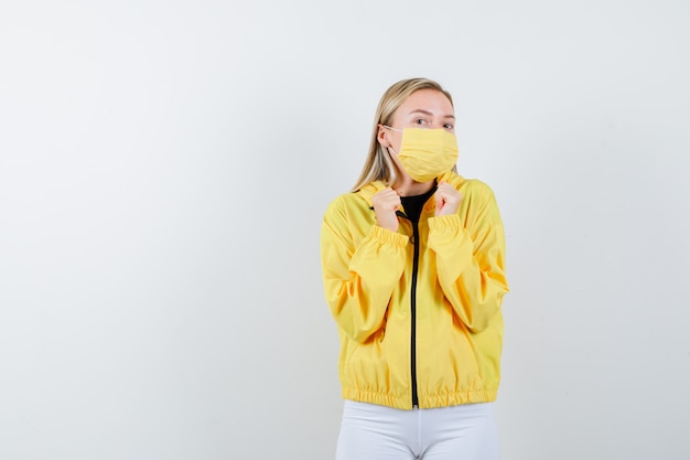 Portrait de jeune femme montrant le geste gagnant en veste, pantalon, masque et à la vue de face chanceux