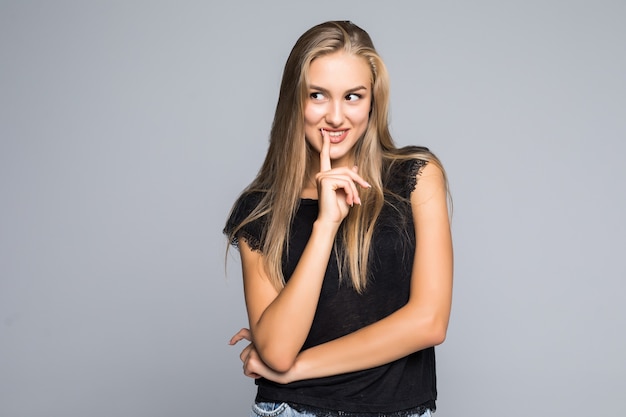 Portrait de jeune femme montrant le geste de la pensée avec le doigt à la lèvre, la bouche ouverte, sur fond gris