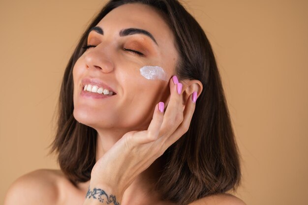 Portrait d'une jeune femme sur un mur beige avec un maquillage naturel chaud et une peau propre et lisse, applique de la crème sur son visage