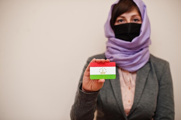 Portrait d'une jeune femme musulmane portant des vêtements de cérémonie protégeant le masque facial et le foulard hijab tenant la carte du drapeau du Tadjikistan sur fond isolé Concept de pays Coronavirus