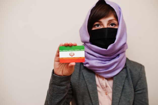 Portrait d'une jeune femme musulmane portant des vêtements de cérémonie protégeant le masque facial et le foulard hijab tenant la carte du drapeau de l'Iran sur fond isolé Concept de pays Coronavirus