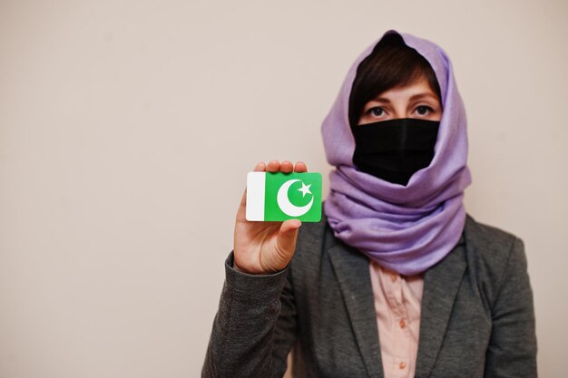 Portrait d'une jeune femme musulmane portant des vêtements de cérémonie protégeant le masque facial et le foulard hijab tenant la carte du drapeau pakistanais sur fond isolé Concept de pays Coronavirus