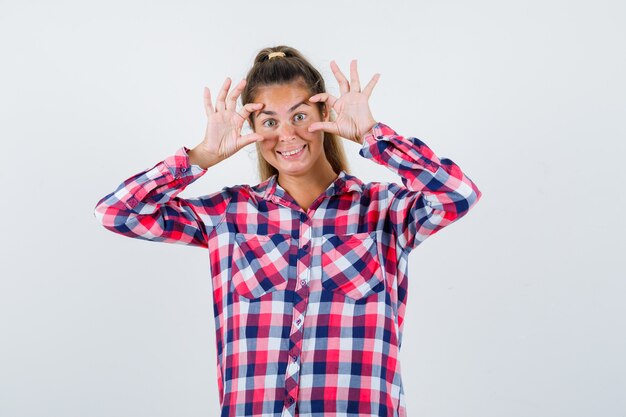 Portrait de jeune femme ouvrant les yeux avec les doigts en chemise à carreaux et à la joyeuse vue de face