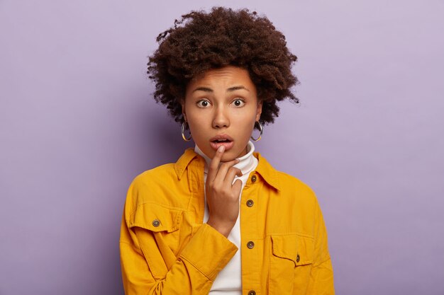 Portrait de jeune femme perplexe nerveuse touche la lèvre inférieure, a une coiffure afro bouclée, une expression anxieuse, porte une veste jaune à la mode et des boucles d'oreilles, des modèles intérieurs sur fond violet