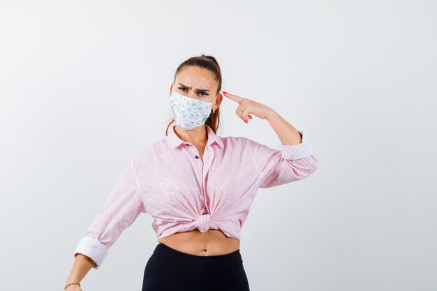 Portrait de jeune femme pointant sur sa tête en chemise, pantalon, masque et à la vue de face pensive