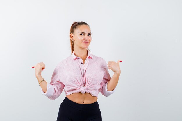 Portrait de jeune femme pointant vers l'arrière avec les pouces en chemise, pantalon et à la vue de face curieuse