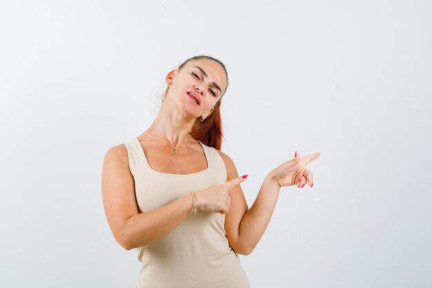 Portrait de jeune femme pointant vers le côté droit en débardeur beige et à la vue de face heureuse