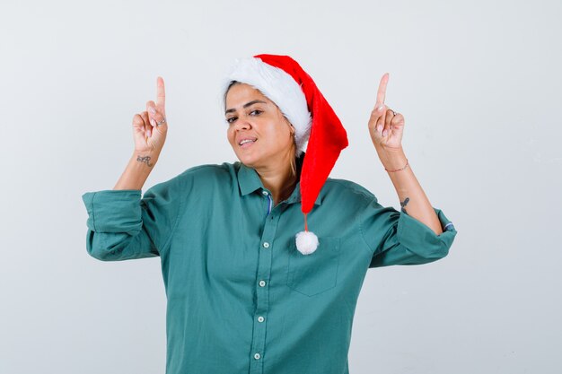 Portrait de jeune femme pointant vers le haut en chemise, bonnet de Noel et à la vue de face confiant