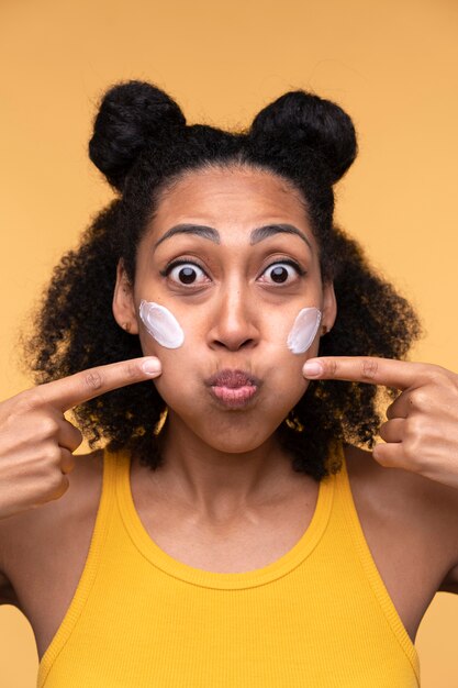 Portrait d'une jeune femme portant une crème hydratante sur son visage et pointant du doigt ses joues enflées