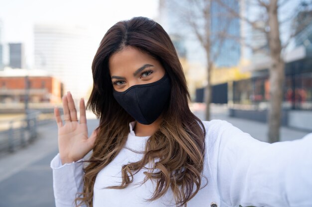 Portrait de jeune femme portant un masque facial et prenant des selfies tout en agitant la main pour dire bonjour à l'extérieur. Concept urbain. Nouveau concept de mode de vie normal.