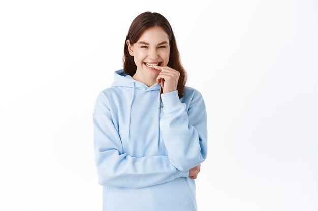 Portrait d'une jeune femme positive et mignonne en sweat à capuche, souriante et riant, touchant les lèvres avec un sourire blanc joyeux, debout contre le mur du studio