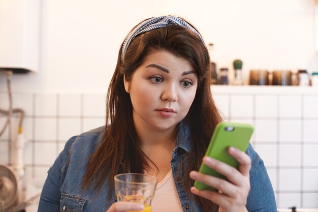 Portrait d'une jeune femme potelée surprise, choquée, buvant du jus de désintoxication d'agrumes maison, à l'aide d'un téléphone intelligent, étonnée par des nouvelles inattendues. Personnes, mode de vie sain et concept technologique