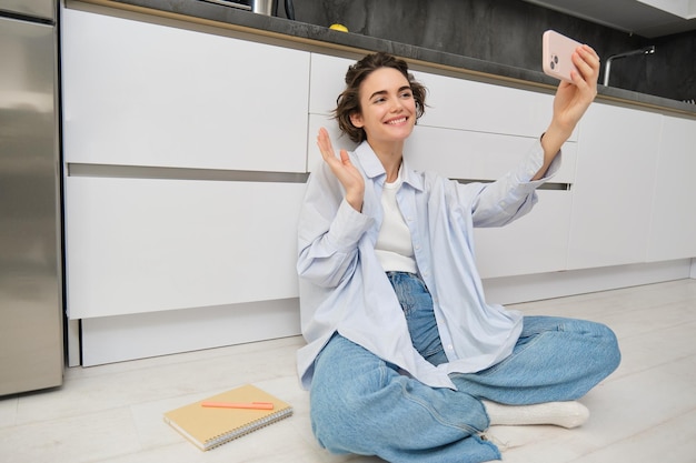 Photo gratuite portrait d'une jeune femme qui discute vidéo enregistre un vlog alors qu'elle est assise sur le sol de la cuisine, une jolie fille prend un selfie