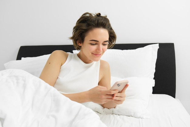 Photo gratuite portrait d'une jeune femme qui se réveille dans son lit en utilisant un smartphone éteindre l'alarme sur le téléphone portable couché dans