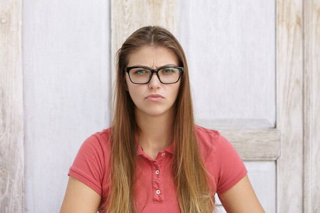 Portrait de jeune femme de race blanche avec de longs cheveux lâches portant des lunettes élégantes et un polo ayant regard bouleversé et malheureux, fronçant les sourcils et faisant le visage ironique, assis contre un mur en bois