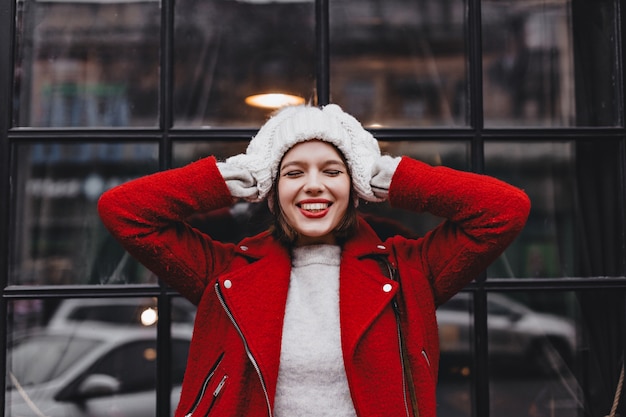 Portrait de jeune femme avec rouge à lèvres et sourire blanc comme neige, vêtue d'un bonnet tricoté blanc et mitaines et veste en laine contre la fenêtre dans un cadre noir.